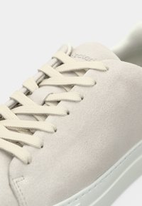 Gros plan d'une chaussure de sport en daim beige avec des lacets crème et une semelle en caoutchouc blanche, montrant des coutures détaillées et la texture.