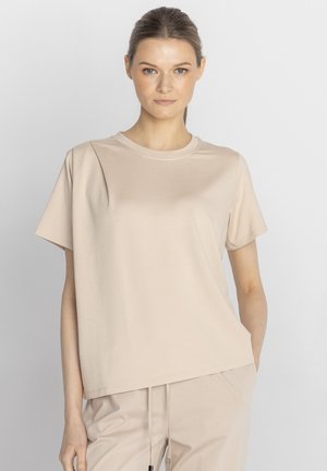 Kobieta o jasnobrązowych włosach nosi beżowy t-shirt z krótkim rękawem oraz pasujące spodnie, stoi na jednolitym jasnoszarym tle.