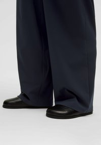 Wide, navy pants made of smooth fabric, paired with black slip-on shoes featuring rounded toes and a matte finish.