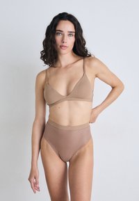 Slušalica i bikinijski donji dio visoko strukirani u mat, svijetlosmeđem materijalu. Slušalica ima tanke naramenice i oblik trokuta; bikini ima teksturirani uzorak pruga.