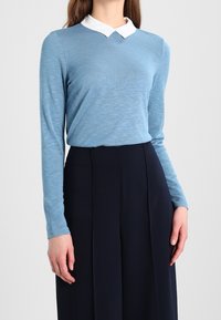 Ljusblå långärmad topp med vit krage, med design i v-form. Tyget ser mjukt ut med en något texturerad yta.