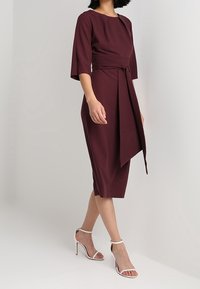 Robe bordeaux avec un corsage ajusté, des manches trois-quarts et une ceinture à la taille. Fabriquée en tissu lisse, longueur au genou, portée avec des sandales à talons à brides.