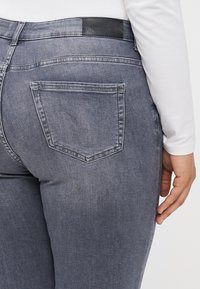 Calças de ganga cinzentas com cintura de altura média, apresentando um acesso em pele preta, um único bolso traseiro e textura suave.