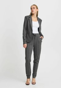 Szürke csíkos blézer, hozzá illő nadrággal, húzózsinórral és slim fit szabással. Fehér toppal és nyitott orrú szandálokkal viselve.