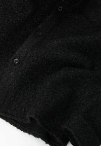 Sort tekstureret stof med en blød, fuzzy overflade og synlige knapper. Har et afslappet, draperet design med en let frynset kant.