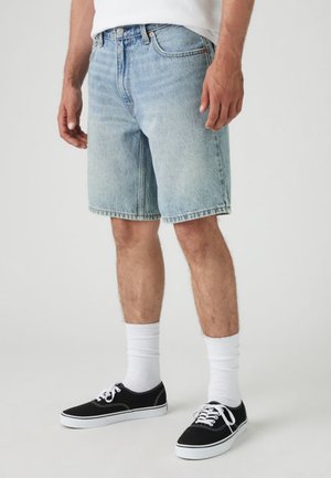 Uomo che indossa pantaloncini di jeans azzurri, calzini bianchi e sneakers nere basse in piedi contro uno sfondo semplice.