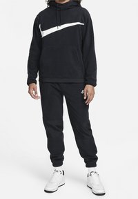 Černá fleecová mikina s kapucí a bílým logem Nike, přední klokaní kapsa, doplněná odpovídajícími tepláky s kontrastními panelami z látky.