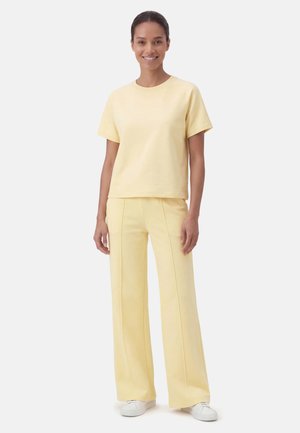 Hymyilevä nainen, joka on pukeutunut vaaleankeltaiseen lyhythihaiseen paitaan ja leveälahkeisiin housuihin valkoiset lenkkarit jalassa, seisoo vaaleanharmaata taustaa vasten.