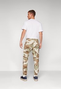 Spodnie cargo w beżowo-zielono-brązowy wzór camouflage, z licznymi kieszeniami, elastycznymi mankietami i luźnym krojem, w połączeniu z białą koszulką.
