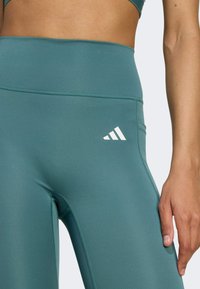Teal athletic leggings made of stretch fabric with a high waistband, featuring white Adidas logo on the left thigh and flat seams.