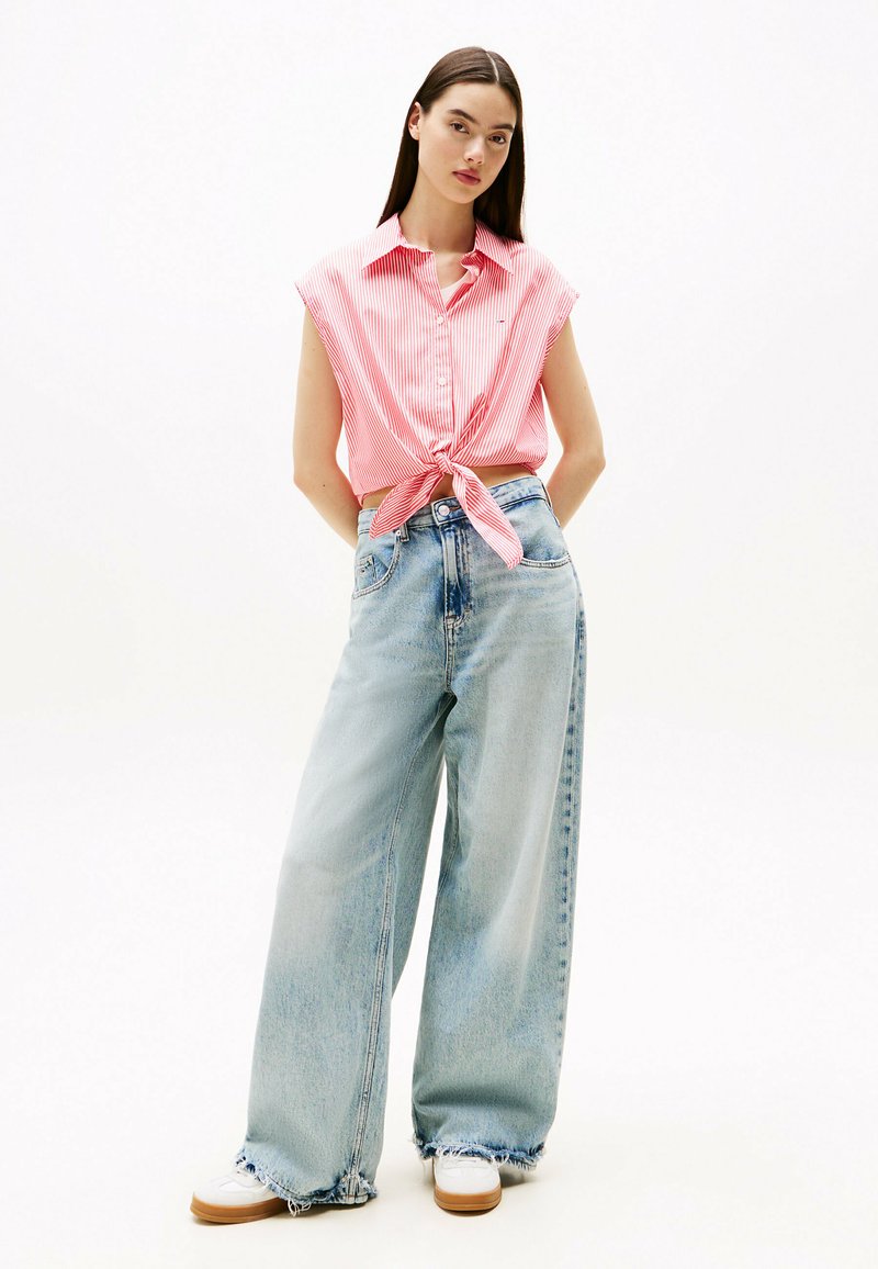 Chemise à rayures rouge et blanche à manches courtes nouée à la taille, associée à un jean léger à jambes larges bleu clair et des baskets blanches avec une semelle texturée.