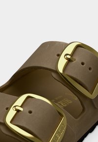 Καφέ λουράκι σανδάλου με δύο μεγάλα λαμπερά χρυσά αγκράφια και εμφανείς ραφές σε λευκό φόντο.