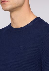 Granatowy sweter wykonany z dzianiny z okrągłym dekoltem, strukturalnym materiałem oraz subtelnie umieszczonym logo na piersi. Miękka tekstura i dopasowany krój.