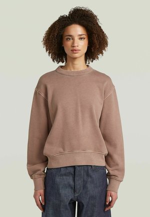 Donna con capelli ricci che indossa una felpa marrone larga e jeans blu scuro, in piedi davanti a uno sfondo chiaro e semplice.