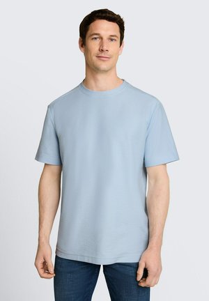 Mężczyzna o krótkich ciemnych włosach, ubrany w jasnoniebieską koszulkę z krótkim rękawem i ciemnoniebieskie jeansy, stojący na tle jednolitego jasnego tła.