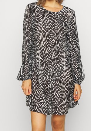 Frau trägt ein langärmliges Kleid mit schwarz-weißem Zebramuster und rundem Ausschnitt, steht vor einem einfarbigen weißen Hintergrund.