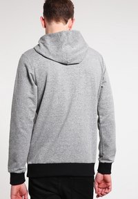 Man die een lichtgrijze capuchontrui draagt met zwarte boorden en zoom, gezien van achteren tegen een effen achtergrond.