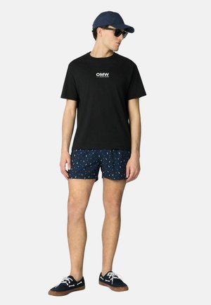 Uomo con berretto nero, occhiali da sole, t-shirt nera con testo "OMW", pantaloncini blu navy a fantasia e sneakers blu navy con lacci bianchi, in piedi di profilo.