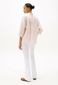 Camisa de lino rosa claro con un pliegue en la parte posterior, mangas enrolladas y un cuello, combinada con pantalones acampanados blancos y mocasines marrones.