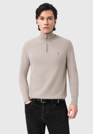 Suéter beige claro con cremallera, cuello alto, textura de punto y pequeño logo bordado en el pecho. Combinado con jeans negros.