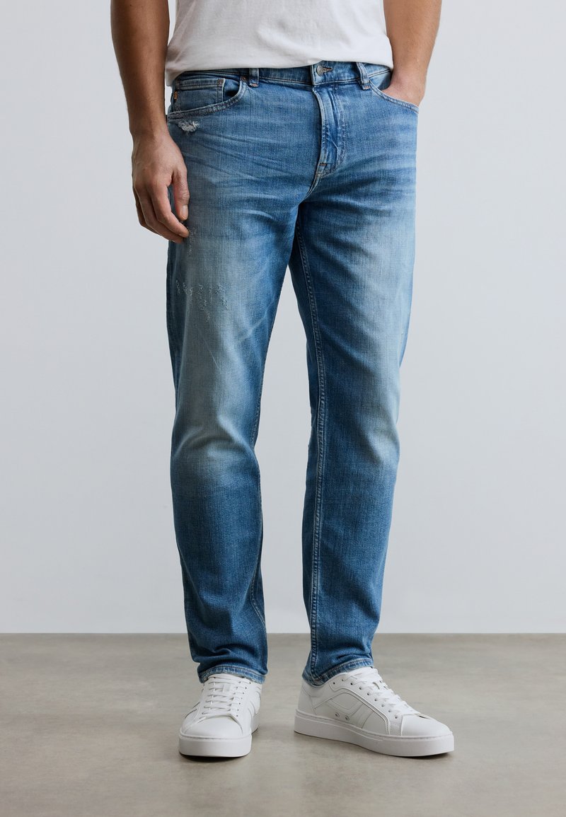 Φωτεινό μπλε τζιν παντελόνι με στενή εφαρμογή, ξεβαμμένες λεπτομέρειες και μικρές φθορές. Συνδυάζεται με λευκά αθλητικά παπούτσια.