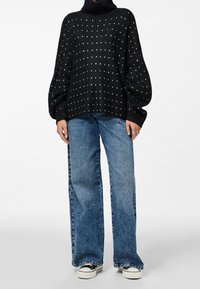 Svart stickad tröja med hög krage och texturerat mönster; prydnad med silverprickar. Kombinerad med vida blå jeans och svarta sneakers.