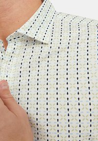 Chemise à manches courtes en tissu léger avec une base blanche, présentant un motif floral répétitif en vert, bleu et marine. Détails de col boutonné visibles.