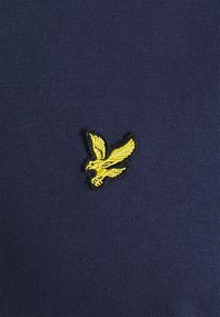 Le tissu marine présente un motif d'aigle jaune brodé, entouré de noir, positionné sur le côté gauche. Texture lisse, design décontracté.