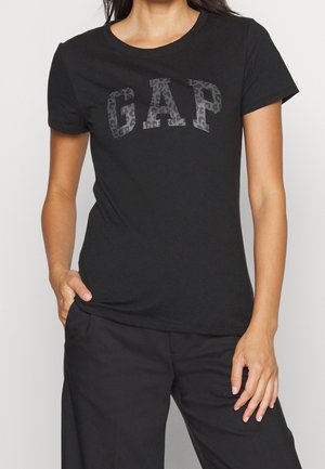 Frau trägt ein eng anliegendes schwarzes T-Shirt mit kurzen Ärmeln und dem Schriftzug "GAP" in Leoprint-Muster auf der Brust.