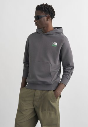 Man draagt een donkergrijze The North Face hoodie, een zonnebril en olijfgroene broek, met handen in de zakken, kijkt naar links tegen een effen achtergrond.