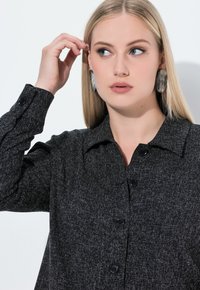 Zwarte button-up shirt met een gestructureerde stof, voorzien van een kraag en lange mouwen. Model draagt grote ovale oorbellen, zachte verlichting, neutrale achtergrond.