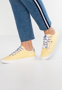 Yellow canvas sneakers with white rubber soles. Features silver heel accents and multicolored laces with brand labels. Blue jeans with black stripes.