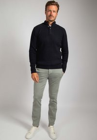 Man staat en draagt een zwarte kabelgebreide trui, lichtgrijze broek en witte sneakers tegen een effen grijze achtergrond.