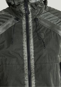 Ciemnozielona kurtka z kapturem, z kontrastującymi szarymi akcentami, z zamkiem błyskawicznym i regulowanymi sznurkami przy kołnierzu i dolnym brzegu.