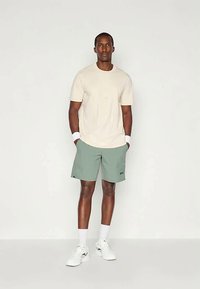 Kreemikatane T-särk, lõdva lõikega; rohelised lühikesed püksid logoga üksikasjal; valged sportlikud kingad; modell seisab käed taskus.