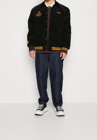 Czarna bomberka z sztruksu z pomarańczowym haftem, paskowanymi mankietami, zapinana na guziki, w połączeniu z ciemnymi dżinsowymi spodniami i czarnymi sportowymi butami.