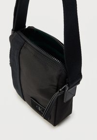Μαύρη τσάντα ώμου από νάιλον με φερμουάρ, υφασμένο πλαϊνό πάνελ και μοτίβο δερμάτινης ετικέτας. Μαλακό λουρί για ρυθμιζόμενη εφαρμογή.