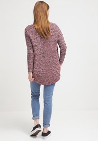 Žena s dlouhými vlasy, oblečená v růžovém a šedém pleteném svetr, modrých zkrácených džínách a černých teniskách, stojící zády k bílé pozadí.