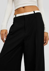 Schwarze weit geschnittene Hose mit glatter Textur und kontrastierendem weißen Bund. Verfügt über seitliche Taschen und einen maßgeschneiderten Schnitt.