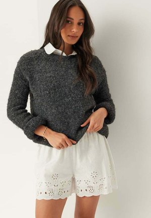 Femme portant un pull texturé gris foncé par-dessus une chemise blanche à col et un short blanc brodé de fleurs, debout contre un mur clair.