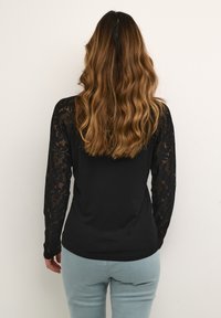 Černá dlouhá halenka s krajkovými rukávy, spárovaná se světle modrými kalhotami. Halenka má hladkou texturu a přiléhavý střih.