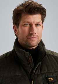 Homme aux cheveux bruns courts et à la barbe de trois jours, portant une veste zippée vert foncé avec le logo PME Legend sur la poitrine et le col.