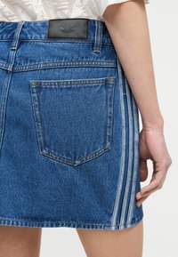 Τζιν φούστα με πίσω τσέπη και πάτς με το λογότυπο της Adidas, που διαθέτει τρεις λευκές ρίγες στην πλευρά, φορεμένη με λευκή υφασμάτινη μπλούζα.