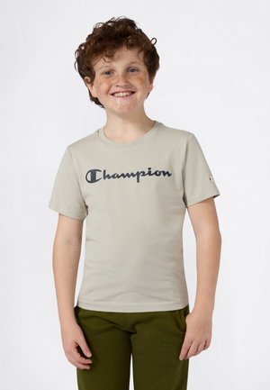 Gaiša bēša kokvilnas T-kreklā ar tumši zilu "Champion" logo priekšpusē; īsas piedurknes un apaļš kakls.