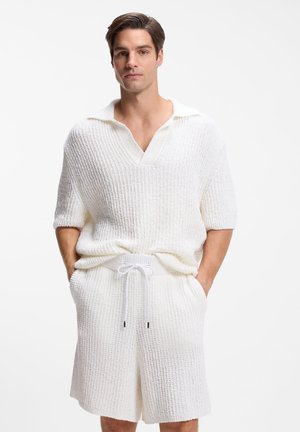 Hombre con suéter de manga corta de punto texturizado blanco y pantalones cortos de punto a juego, con las manos en los bolsillos, de pie frente a un fondo blanco liso.