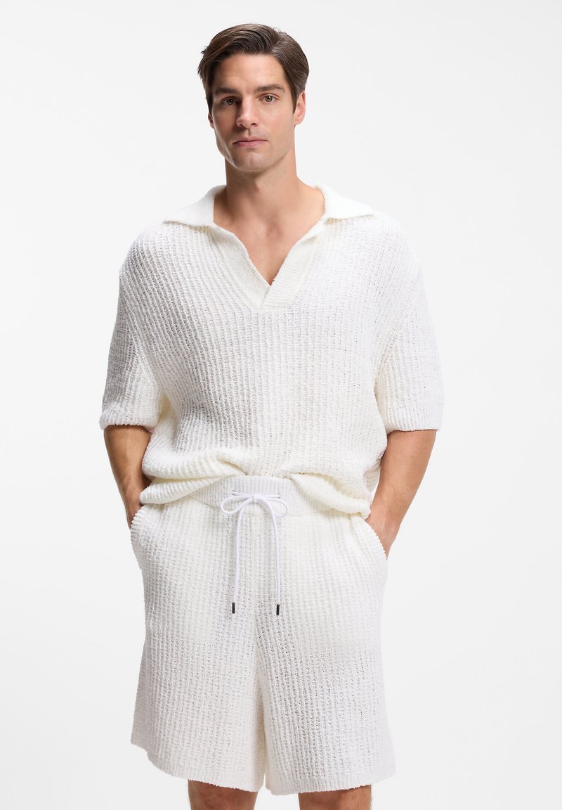 Άνδρας που φοράει λευκό ύφασμα πλεκτό πουλόβερ με κοντά μανίκια και αντίστοιχα πλεκτά σορτς, με τα χέρια στις τσέπες, στέκεται μπροστά σε ένα απλό λευκό φόντο.
