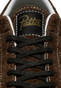 Brown suede sneaker with black laces. Features a white stitched trim and a black patch with gold "Patta" logo. Close-up of the tongue.