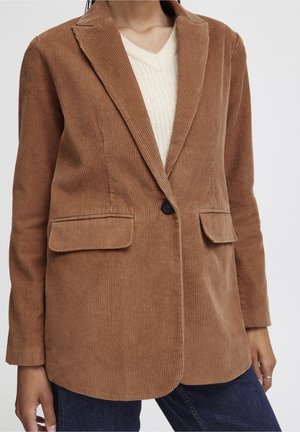 Femme portant un blazer en velours côtelé marron à un bouton sur un pull crème à col en V et un jean bleu foncé.