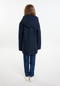 Sötétkék kapucnis dzseki strukturált felülettel, hosszú ujjakkal és egyenes szabással, kék farmernadrággal és fehér sportcipővel párosítva.