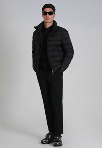 Doudoune noire avec un col montant, chemise noire en dessous, pantalon noir ajusté et chaussures de sport noires avec une finition texturée.